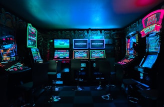 Dimly lit gaming arcade featuring a variety of brightly illuminated slot machines and video gaming screens. Neon signs and colorful displays create a vibrant atmosphere. The arrangement of gaming machines suggests an inviting space for entertainment, with chairs positioned for players.